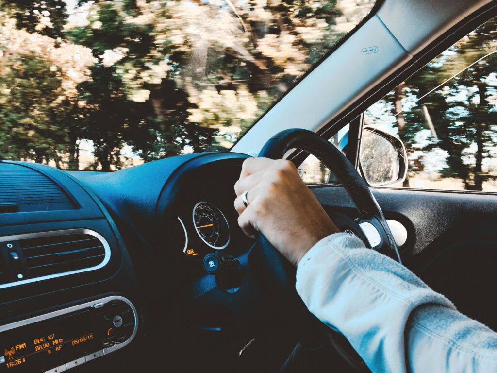 pexels-photo-1392621-1392621 Driver's hand on steering wheel of a modern car during a daytime drive through scenic outdoors.